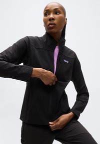 Γυναίκα με κοντά μαλλιά που φοράει μαύρο μπουφάν με φερμουάρ πάνω από ένα μωβ πουκάμισο, τραβώντας ελαφρώς το φερμουάρ του μπουφάν, μπροστά σε απλό φόντο.