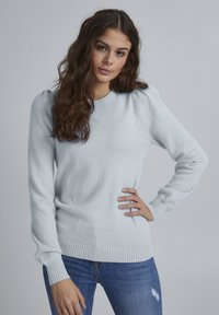 Jasnoniebieski sweter wykonany z miękkiego, teksturowanego materiału. Posiada ściągacz przy dekolcie i mankietach oraz lekko bufiaste rękawy. Noszony z niebieskimi dżinsami.