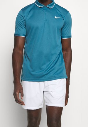 Mann trägt ein türkisfarbenes Polohemd mit kurzen Ärmeln und weißem Rand sowie Nike-Logo, kombiniert mit weißen Sportshorts, steht vor einem einfarbigen Hintergrund.