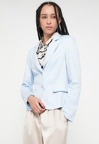 Blazer azul claro a medida con solapas con muesca, cierre de un solo botón y dos bolsillos laterales, usado sobre una blusa estampada y pantalones claros.
