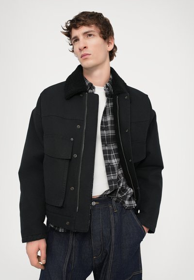 Black jacket with a textured faux shearling collar, front pockets, worn over a checkered flannel shirt and white undershirt. Dark jeans.