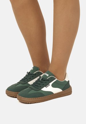 Persona con zapatillas bajas verdes y blancas con suelas marrones, de pie sobre un fondo claro y liso.