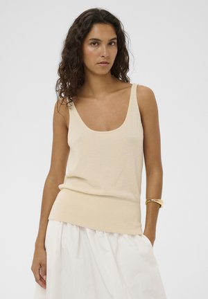 Vrouw met krullend donker haar, gekleed in een crèmekleurige mouwloze gebreide top en een witte rok, staand met één hand in haar zak tegen een effen achtergrond.