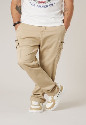 Homme portant un pantalon cargo beige avec poches zippées et des baskets beige et blanches, debout les jambes croisées et les mains dans les poches.