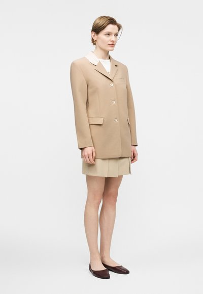 Γυναίκα που φοράει μπεζ σακάκι πάνω από λευκό τοπ και σύντομη μπεζ πλισέ φούστα, συνδυασμένη με σκούρα καφέ επίπεδα παπούτσια, στέκεται μπροστά σε λευκό φόντο.