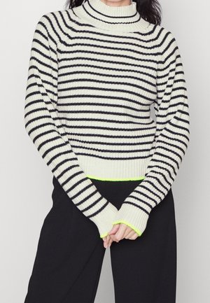 Femme portant un pull à col roulé blanc côtelé avec des rayures horizontales noires et un bord néon jaune aux poignets et à l'ourlet, assorti à un pantalon noir.