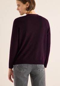 Dunkellila Strickpullover mit langen Ärmeln, rundem Hals, gerippten Bündchen und Saum. Kombiniert mit grauen, strukturierten Jeans aus Sicht der Seite und von hinten.