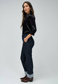 Blusa de satén negra con mangas cortas, combinada con jeans de denim azul oscuro con dobladillos. Botines de tacón marrones complementan el look.