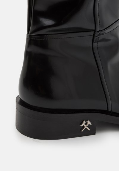 Svart lackläderstövel med en kraftig svart sula och metallisk accent med ett korsat hammare- och yxdessign. Slen yta, elegant form.