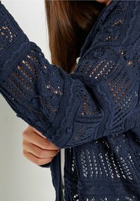 Marineblauer Strickcardigan mit einem offenen Design, strukturierten Mustern und einer lockeren Passform, ausgestattet mit langen, detaillierten Ärmeln.