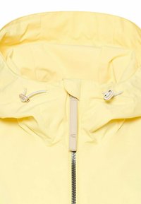 Żółta kurtka z kapturem wyposażonym w regulowane sznurki i centralny zamek błyskawiczny z beżowym uchwytem.