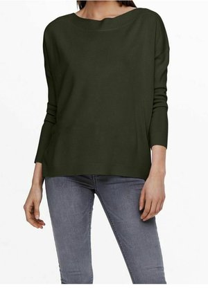 Femme portant un pull ample vert foncé à manches longues avec un large col et un jean gris ajusté, debout les bras détendus.