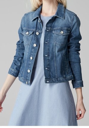 Frau trägt eine mittelblaue Jeansjacke über einem hellblau-weiß gestreiften Kleid vor neutralem grauem Hintergrund.