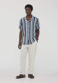 Camicia a maniche corte con bottoni e righe verticali blu e nere, abbinata a pantaloni color crema e sandali neri a infradito.