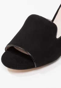 Mule ouvert en daim noir avec une large bride avant et un détail de bordure subtile sur fond blanc.