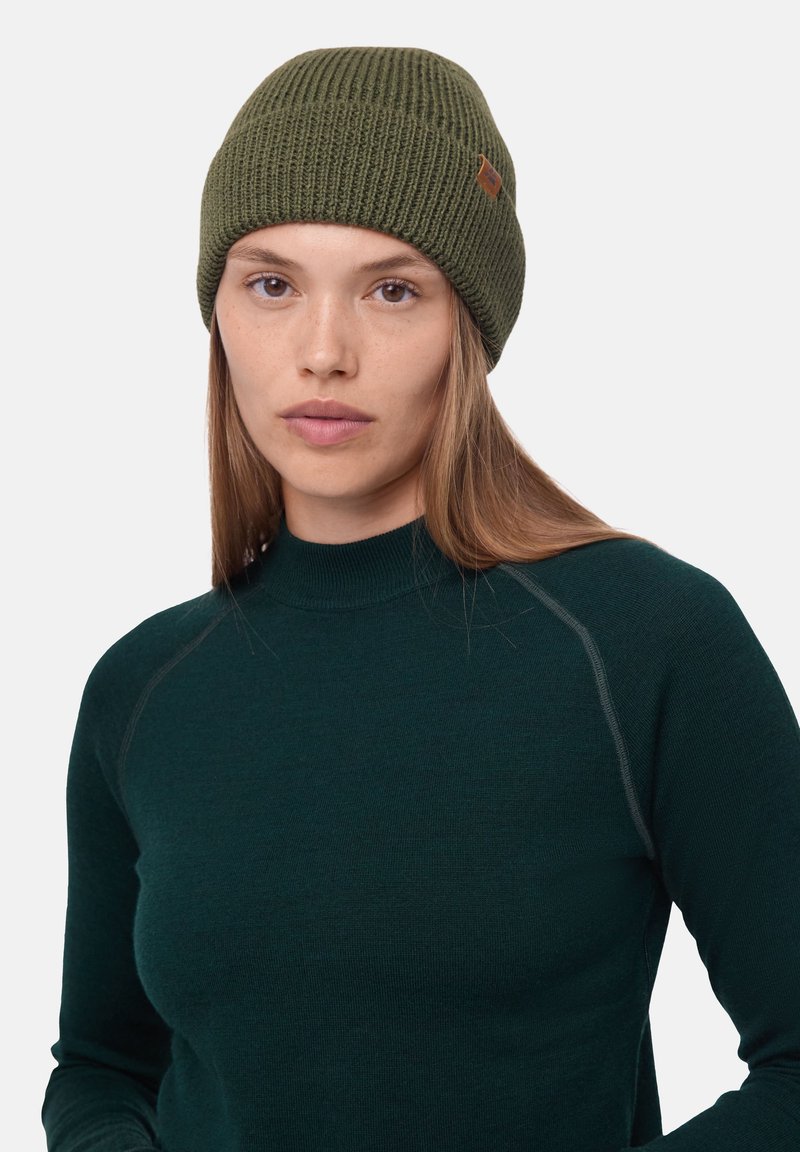 Olive green knit beanie with a ribbed texture and a small leather tag. Worn with a dark green fitted top.