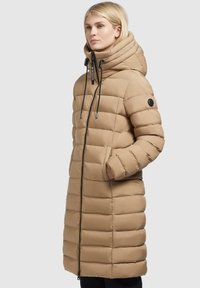 Langer, beigefarbener Steppmantel mit hohem Kragen, Reißverschluss und elastischen Zugbändern. Verfügt über horizontale Steppnähte und Seitentaschen.