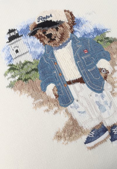 Πλεκτή αρκούδα που φοράει λευκό καπέλο, τζιν μπουφάν, λευκό πουλόβερ, παντελόνι και μπλε παπούτσια, με φόντο φάρο και πρασινάδα.