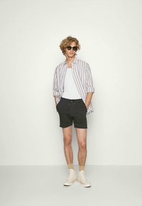 Randig ljusgrå och vit skjorta med knappar, vit linne, svarta shorts och cremefärgade sneakers. Modellen står med händerna i fickorna.
