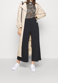 Dlouhý béžový trenčkot přes květinový zelený top, doplněný o černé široké kalhoty a bílé tenisky. Hladká textura, uvolněný střih.