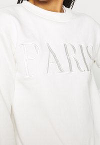 Bílá mikina se stříbrným vyšitým nápisem "PARIS", kulatým výstřihem, žebrovanými manžetami a hladkým bavlněným povrchem.