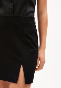 Nahaufnahme einer Person in einem schwarzen Rock mit einem kleinen Schlitz vorne und einem passenden schwarzen Oberteil mit Knöpfen, die vor einem weißen Hintergrund steht.