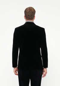 Uomo in piedi di spalle che indossa un blazer di velluto nero e pantaloni neri abbinati su uno sfondo bianco semplice.