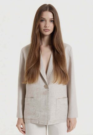 Giovane donna con lunghi capelli castani lisci che indossa un blazer beige chiaro con grandi tasche e pantaloni bianchi su sfondo uniforme.