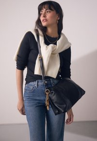 Mustast nahast käekott kuldse kettaktsendiga, kantud kreemja kudumissalliga musta topi peal, kombineeritud siniste teksapükstega.