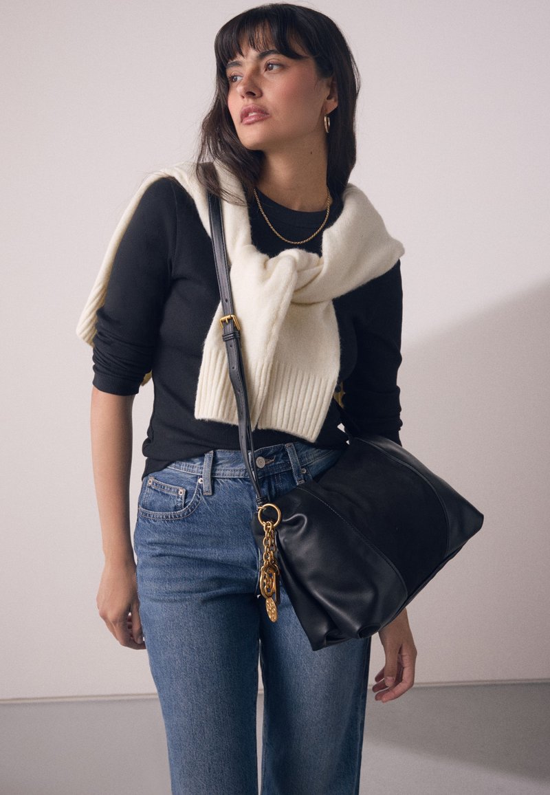 Mustast nahast käekott kuldse kettaktsendiga, kantud kreemja kudumissalliga musta topi peal, kombineeritud siniste teksapükstega.