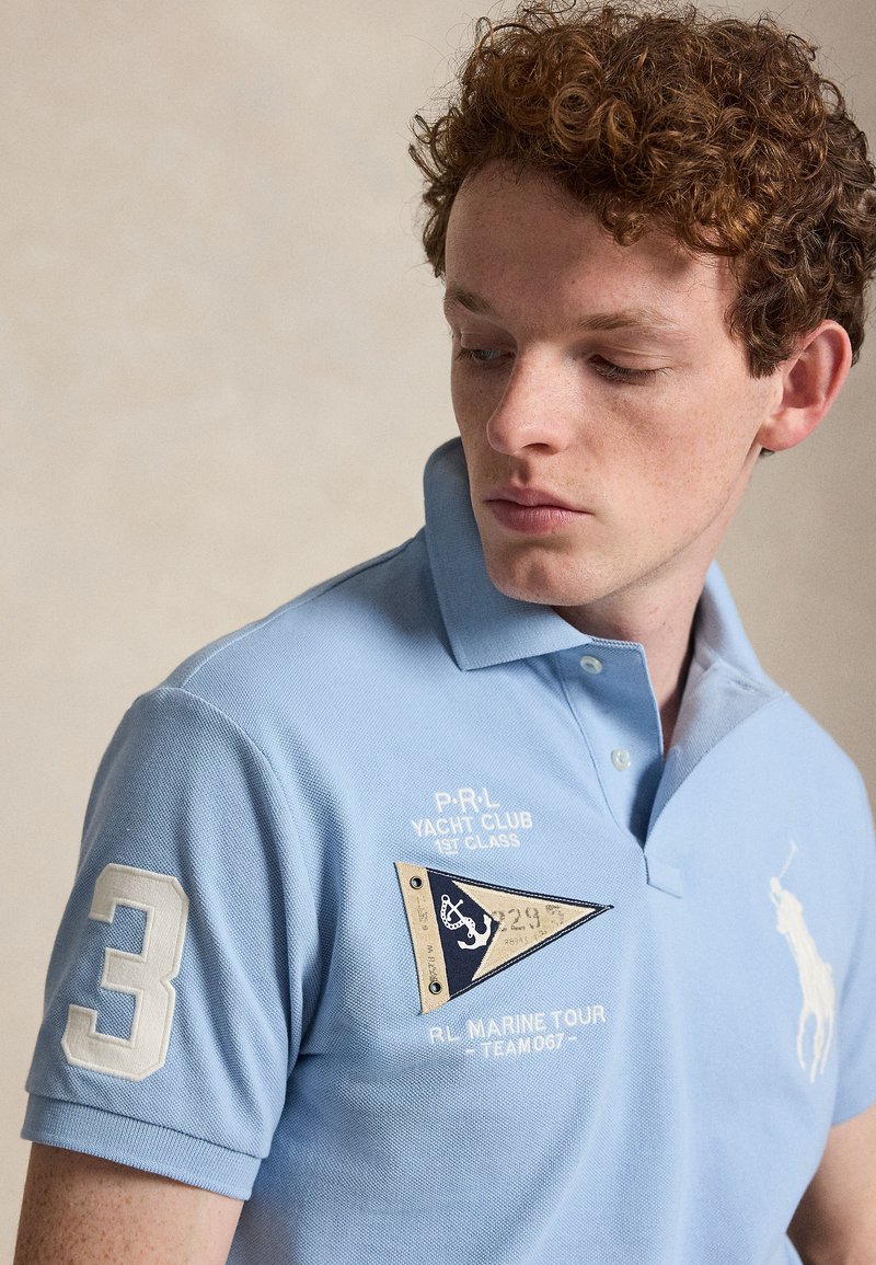 Junger Mann mit lockigem roten Haar, der ein hellblaues Poloshirt mit Yachtclub-Abzeichen und der Nummer 3 am Ärmel trägt.