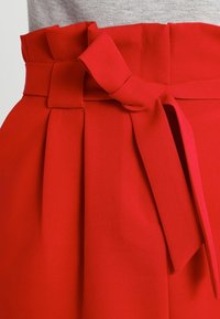 Sukně vysokého pasu jasně červené barvy s plisovaným pasem a zavázaným látkovým opaskem, nošená s šedým topem.