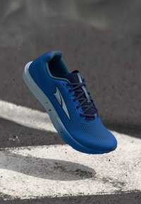 Μπλε αθλητικό παπούτσι με μαύρα κορδόνια και λευκό λογότυπο, αιωρούμενο πάνω από άσφαλτο με λευκές σημάνσεις δρόμου και σκορπισμένα μικρά απορρίμματα.