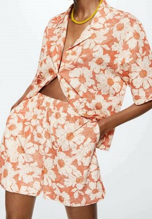 Personne portant une chemise à manches courtes et un short assortis orange et blanc à motifs floraux, les mains dans les poches et un collier de perles jaunes.