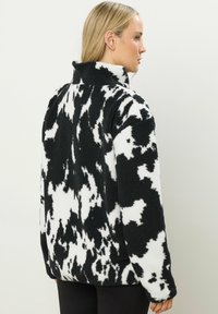 Veste en polaire à motif vache noir et blanc avec un col haut, des épaules arrondies et une surface douce et texturée. Coupe régulière sans quincaillerie visible.