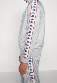Homme portant un survêtement gris clair avec des bandes du logo de la marque en bleu et blanc bordées d’un liseré rouge le long des manches et du pantalon.