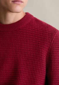 Roter, strukturiert gestrickter Pullover mit einem gerippten Rundhalsausschnitt, der ein Rautenmuster aufweist und aus weichem, mittelschwerem Material besteht. Nahaufnahme.