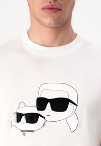 T-shirt in cotone bianco con una grafica in bianco e nero che raffigura un personaggio e un gatto che indossano occhiali da sole neri oversize, con dettagli minimi.