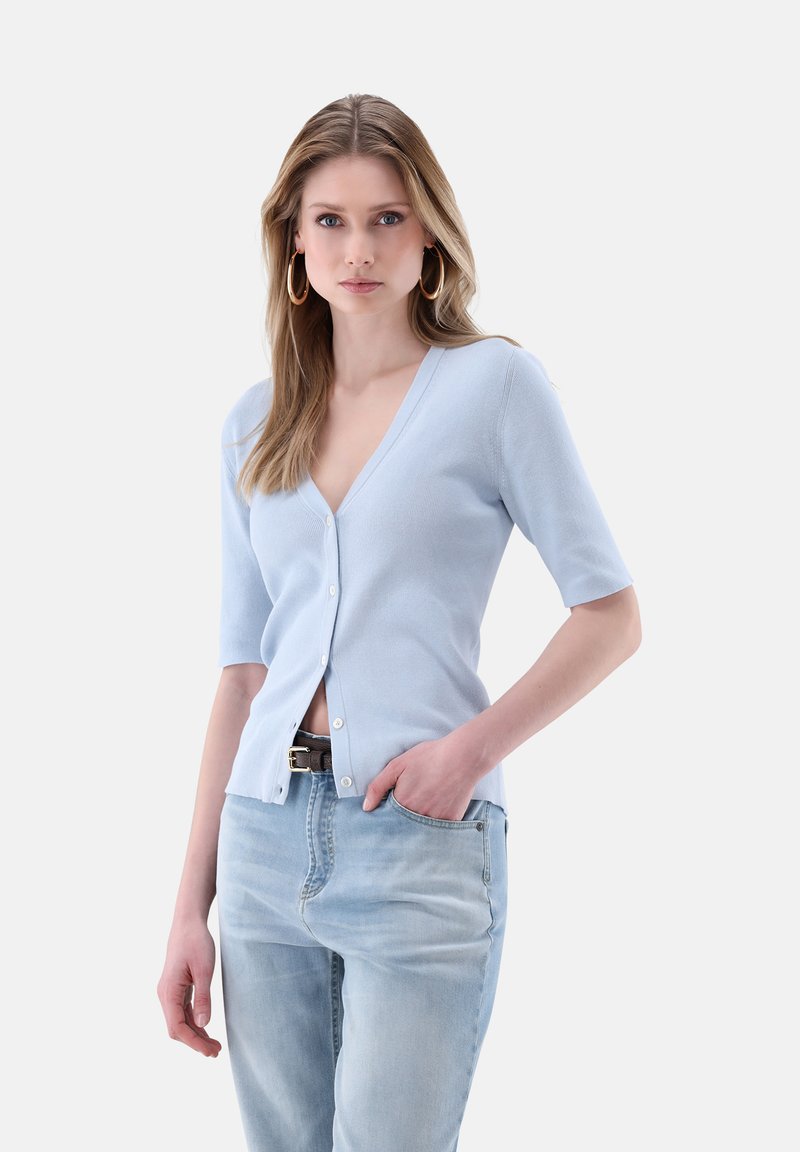 Hellblauer kurzärmeliger Cardigan mit V-Ausschnitt, gerippter Textur und Knopfverschluss, getragen über hellblauen Jeans.