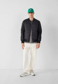 Chaqueta negra acolchada con cierre de cremallera y puños acanalados, combinada con una camisa gris, pantalones blancos y una gorra de béisbol verde.