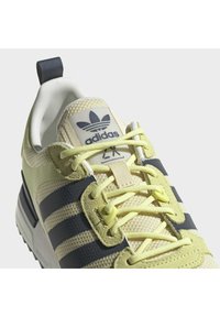 Light yellow athletic shoe with mesh upper, navy stripes, and contrasting laces; includes logo tag on tongue and padded collar for comfort.
