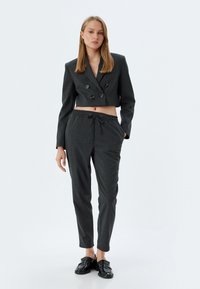 Korte, donkergrijze blazer met dubbele knopen, gecombineerd met donkergrijze, taps toelopende broek met een verstelbare trekkoord in de taille. Zwarte schoenen.