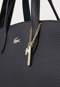 Sac à main en cuir texturé noir avec doubles poignées, accents en matériel doré et logo Lacoste. Comprend un charme et un détail de tirette de fermeture éclair.