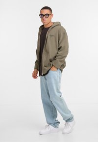 Olivgrön zip-up hoodie över en mörk tröja, kombinerad med ljusblå baggy jeans och vita sneakers. Har fickor och justerbar huva.