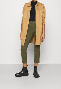 Person som bär en ljusbeige lång knappad skjorta över en svart topp, olivgröna högmidjade avkortade byxor och svarta ankelstövlar, stående på ett grått golv.