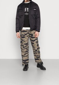 Chaqueta acolchada negra con dos bolsillos frontales sobre un sudadera gráfica negra, combinada con pantalones cargo de camuflaje y botas negras de tobillo.