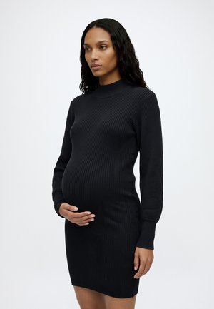 Femme enceinte aux longs cheveux bouclés portant une robe noire côtelée à manches longues, debout devant un fond clair uni.