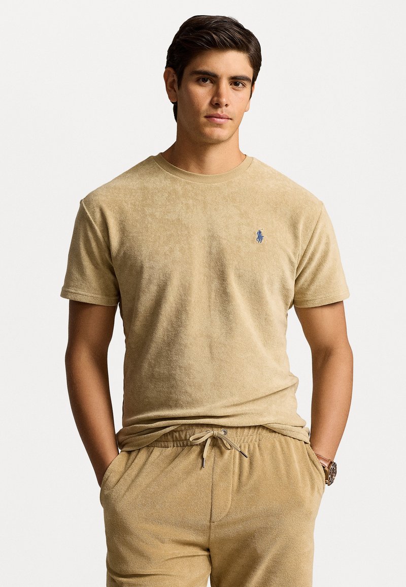 Beige kokvilnas t-krekls ar teksturētu apdari, īsām piedurknēm un zilu izšūtu logotipu kreisajā krūtīs, kopā ar atbilstošām brīvroku biksēm.