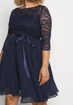 Kvinde iført en mørkeblå kjole med blondekorsage, trekvartærmer og et satinbånd i taljen, stående foran en ensfarvet lys baggrund.