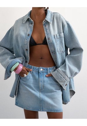 Femme portant une veste en jean bleu clair et une jupe assortie avec un haut de bikini noir et des bracelets colorés au poignet, posant avec les mains dans les poches.